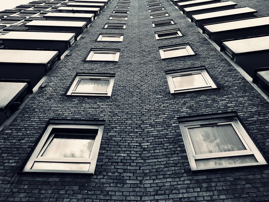 Close-up view of a multi-storey residential building with numerous balconies featuring black metal railings and concrete surfaces. Some balconies display potted plants, outdoor furniture, and laundry